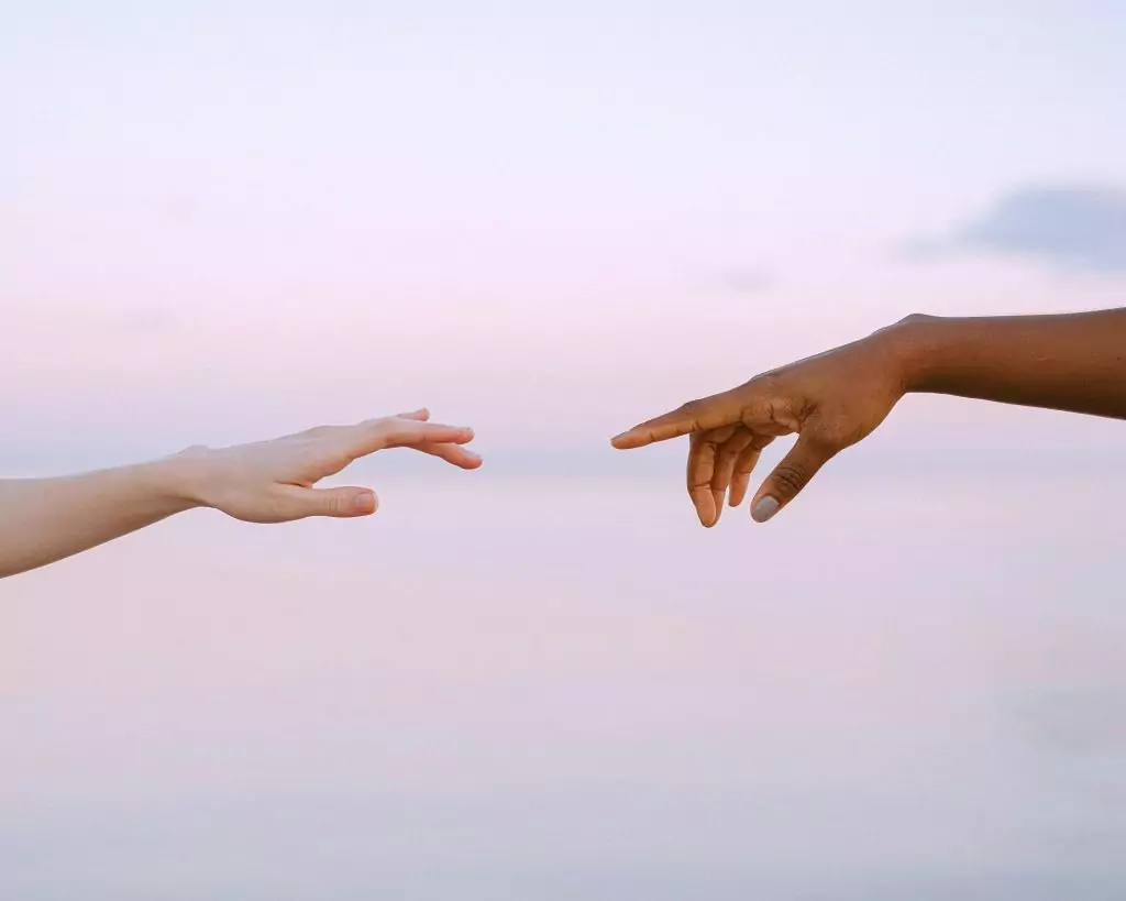 Sinnbild für Premium Begleitung für Erwachsene mit Alexandra Wittek. Zwei Hände reichen jeweils von links und rechts in das Bild rein und sind kurz davor sich mit dem Zeigefinger zu berühren. Der Hintergrund sind rosa Wolken.